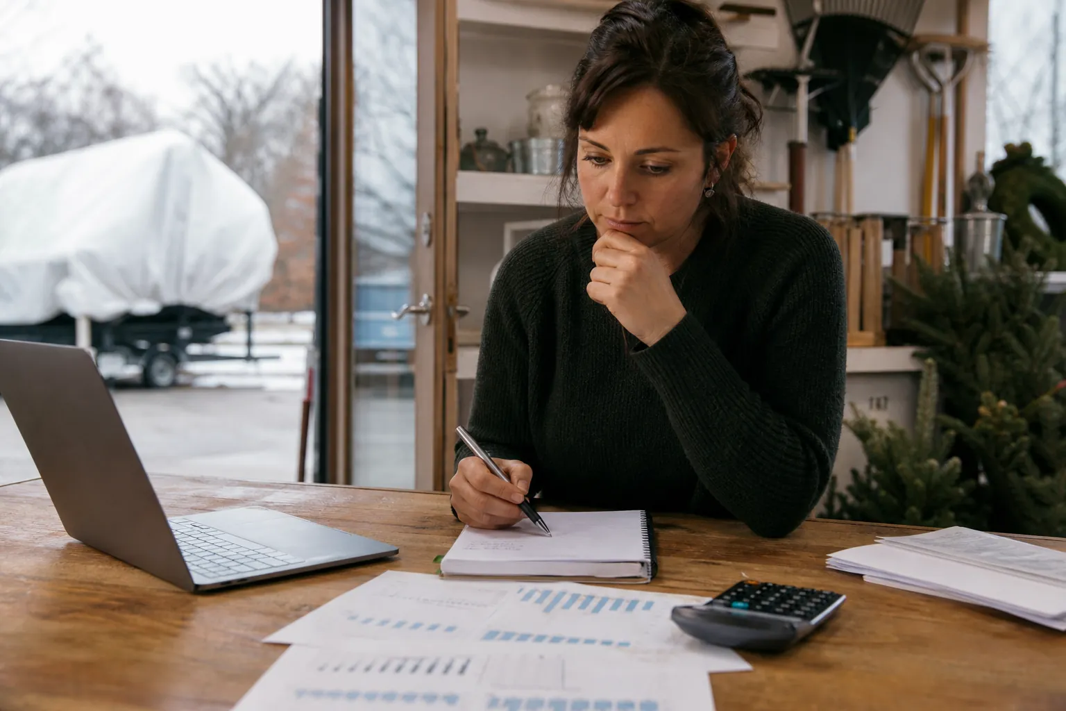 Reviewing Cash Flow During a Seasonal Slow Period Woman reviews financial charts with a laptop, calculator, and notebook beside seasonal greenery in a workshop.