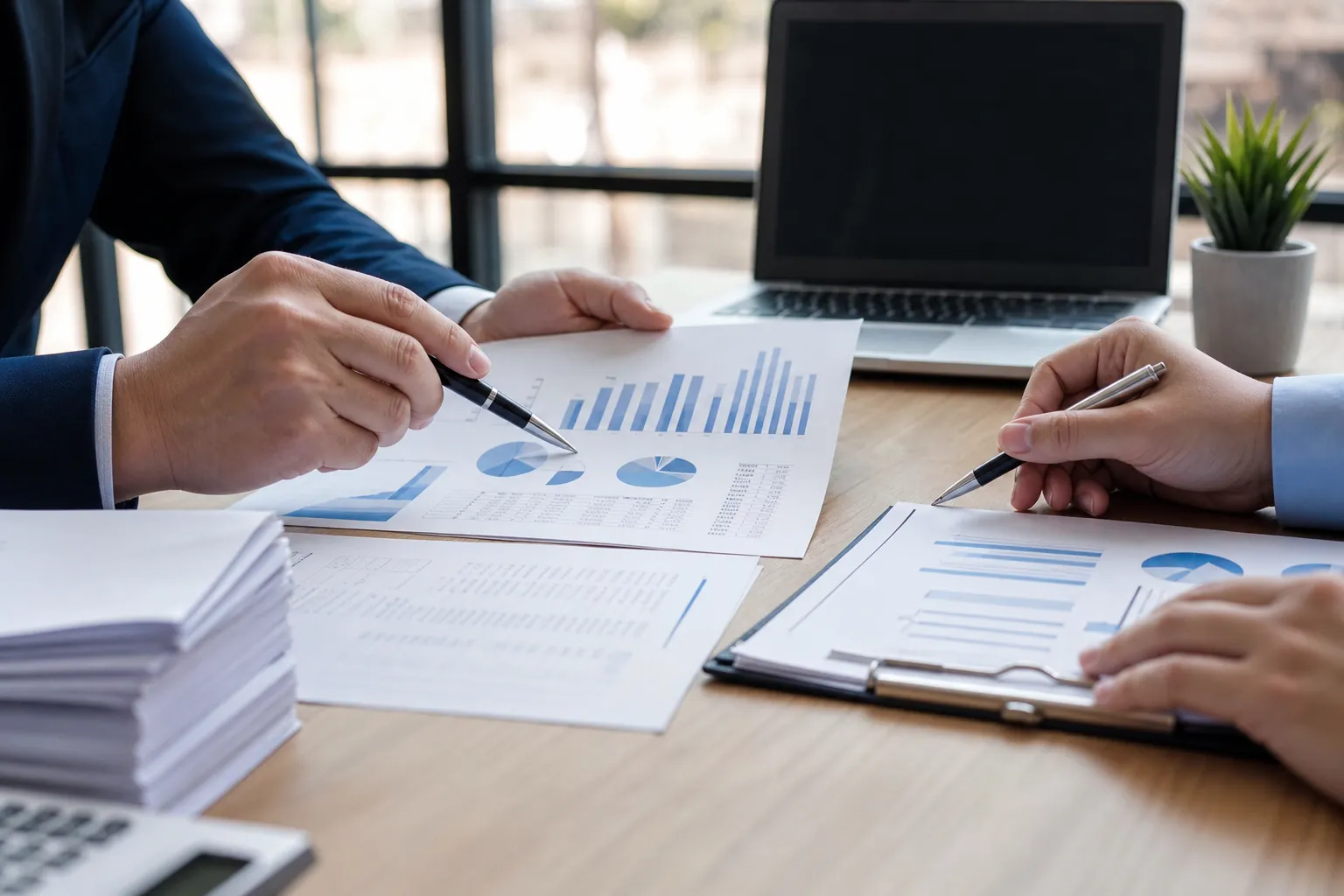 Businesspeople Reviewing Financial Statements and Charts Two businesspeople use pens to review printed financial charts and tables beside a laptop and calculator.