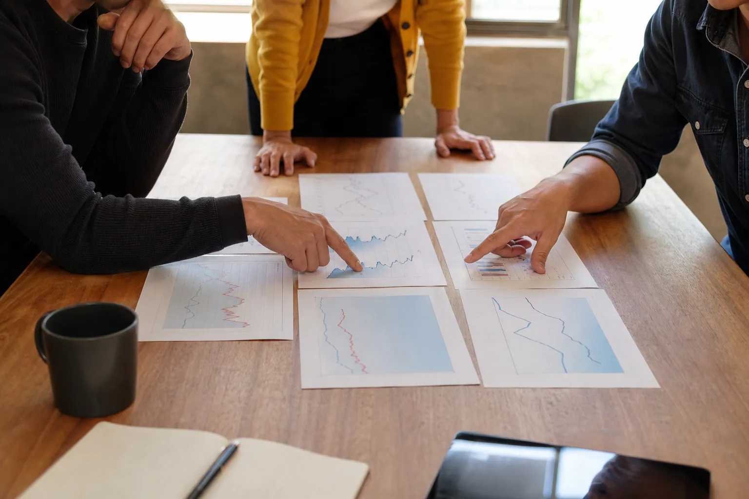 Colleagues reviewing financial forecast charts Three colleagues review and point to printed financial charts spread across a wooden table during a planning meeting.