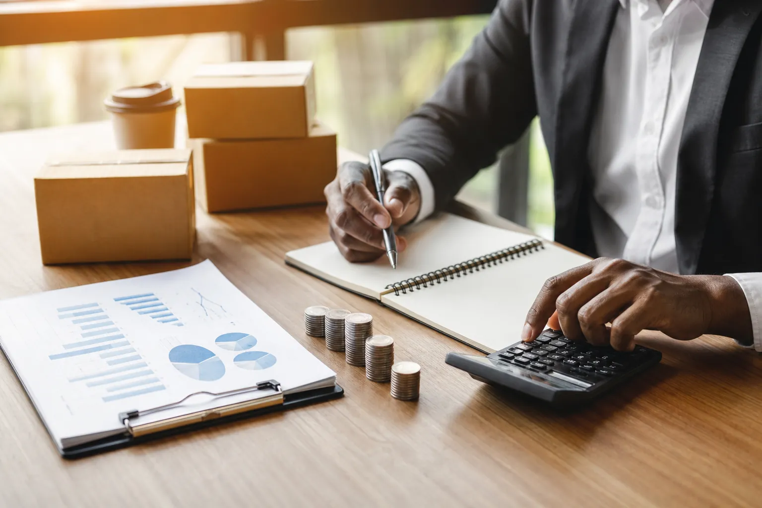 A person in a suit uses a calculator beside an open notebook, stacked coins, financial charts, and small cardboard boxes.